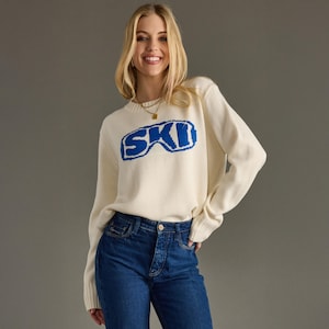 May include: Cream-colored sweater with the word "SKI" in blue, designed to resemble ski goggles. The sweater is paired with blue jeans. The model is smiling and wearing a gold necklace.
