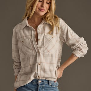 May include: A white and pink plaid flannel shirt with long sleeves and a button-down front. The shirt has two chest pockets and is worn over blue jeans.