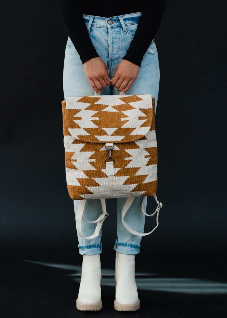 May include: A white and brown geometric patterned backpack with adjustable straps. The backpack is being held by a person wearing blue jeans and white ankle boots.
