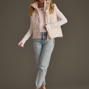 May include: A pink floral print puffer vest with a white long-sleeved shirt and light blue jeans. The model is wearing light blue sandals.