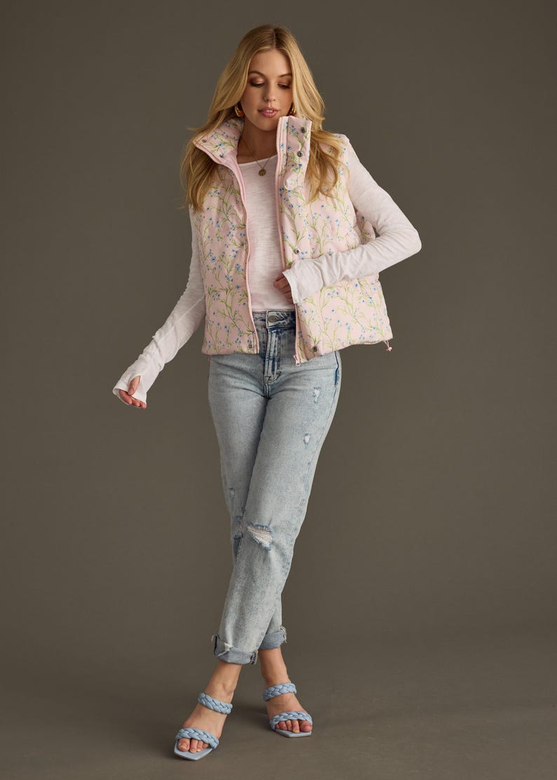 May include: A pink floral print puffer vest with a white long-sleeved shirt and light blue jeans. The model is wearing light blue sandals.