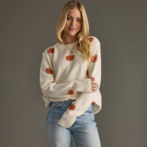 May include: A cream-colored long-sleeve knit sweater featuring a repeating pattern of small, orange pumpkin graphics. The sweater has a ribbed crew neckline and wide ribbed cuffs at the wrists, paired with light blue denim jeans.