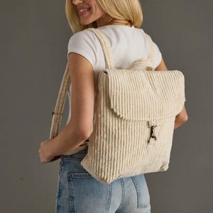 May include: Cream-colored corduroy backpack with vertical ribbing and a flap closure. The backpack has adjustable straps and a metal clasp. The bag is worn over the shoulders.