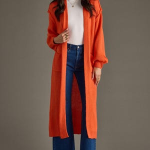 May include: An orange longline cardigan with long sleeves and front pockets. The cardigan is worn over a white top and blue jeans. The model is wearing high-heeled sandals. The cardigan is a stylish outerwear piece.