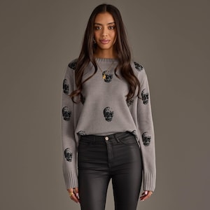 May include: A long-sleeved, crew-neck sweater in a medium gray knit fabric featuring a repeating black skull pattern across the front and sleeves. The garment has ribbed cuffs and a ribbed hem, providing a structured fit for this knit pullover.