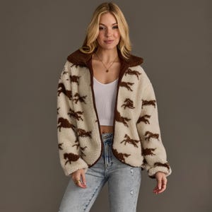 May include: A cream-colored, long-sleeve, zip-up fleece jacket featuring a repeating pattern of brown horses. The jacket has a dark brown collar and trim, a full-length front zipper, and a soft, textured sherpa-like material throughout the garment.