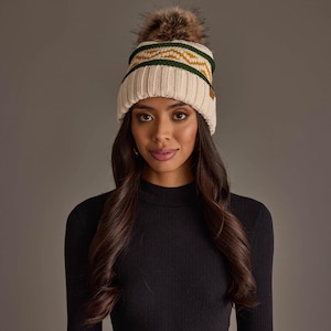 May include: A cream-colored knit beanie featuring a ribbed cuff, a dark green and mustard yellow geometric mountain pattern, and a large, fluffy brown faux fur pom-pom on top. The hat includes a small brown rectangular tag on the side.