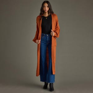 May include: A long, open-front rust-colored knit cardigan with long sleeves and patch pockets, worn over a black short-sleeved top and dark blue denim jeans. The outfit is completed with black ankle boots and a simple gold pendant necklace.