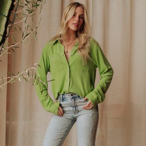 May include: A bright green button-down shirt with two front pockets. The long-sleeved shirt is paired with light-wash blue jeans. The model is wearing a gold necklace. The background is a neutral beige.