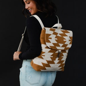 May include: A white and brown geometric patterned backpack with white straps. The backpack is being worn by a person wearing a black long-sleeved shirt and light blue jeans.