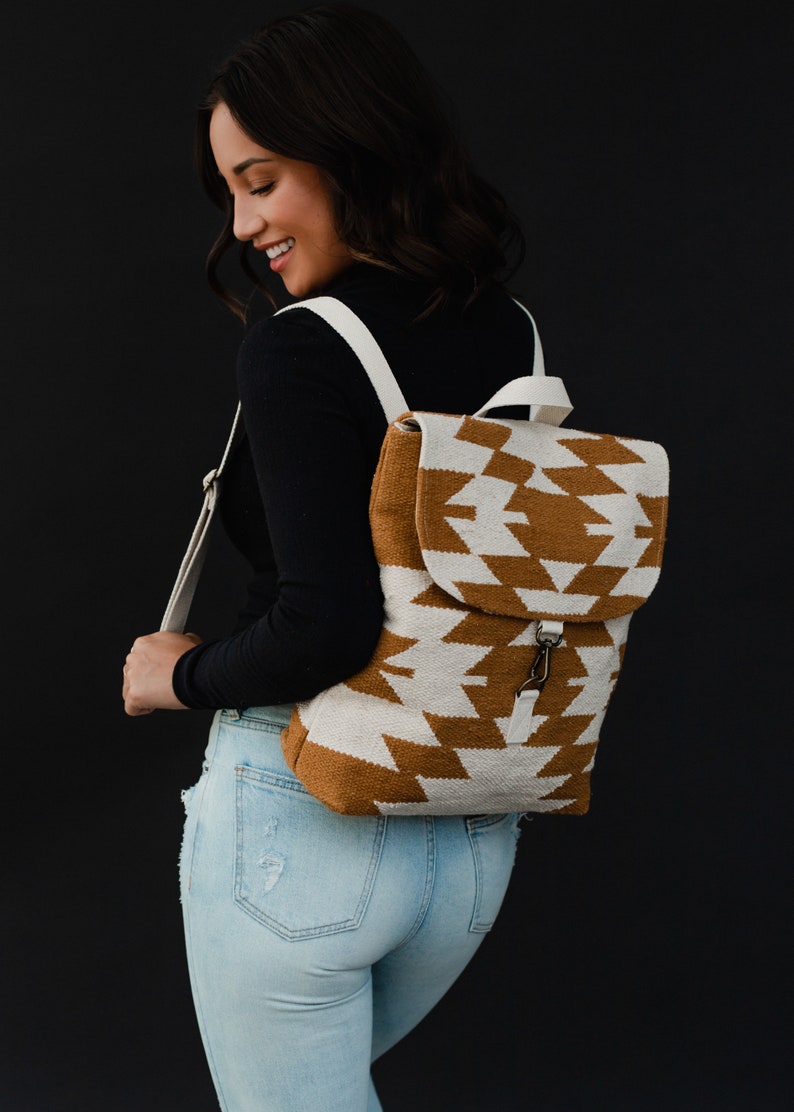 May include: A white and brown geometric patterned backpack with white straps. The backpack is being worn by a person wearing a black long-sleeved shirt and light blue jeans.
