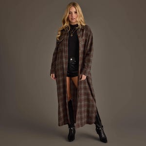 May include: A long-sleeved, floor-length brown plaid shirt dress featuring a button-down front, a classic collar, and two chest pockets. The garment is worn over a black mock-neck top, black shorts, and black leather boots with silver hardware.