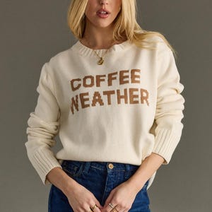 May include: Cream-colored sweater with the words "COFFEE WEATHER" in brown block letters. The sweater has long sleeves and ribbed cuffs. The person is wearing blue jeans and gold rings.