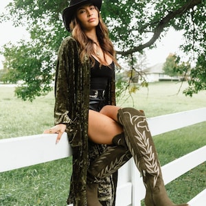 May include: A black cowboy hat, a long olive green velvet duster, a black tank top, black shorts with a silver buckle belt, and olive green knee-high boots featuring white stitched leaf patterns and stacked wooden heels.