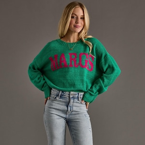 May include: A green sweater with the word "MARGS" in pink across the chest. The sweater has a crew neck and long sleeves. The person in the image is wearing light blue jeans.