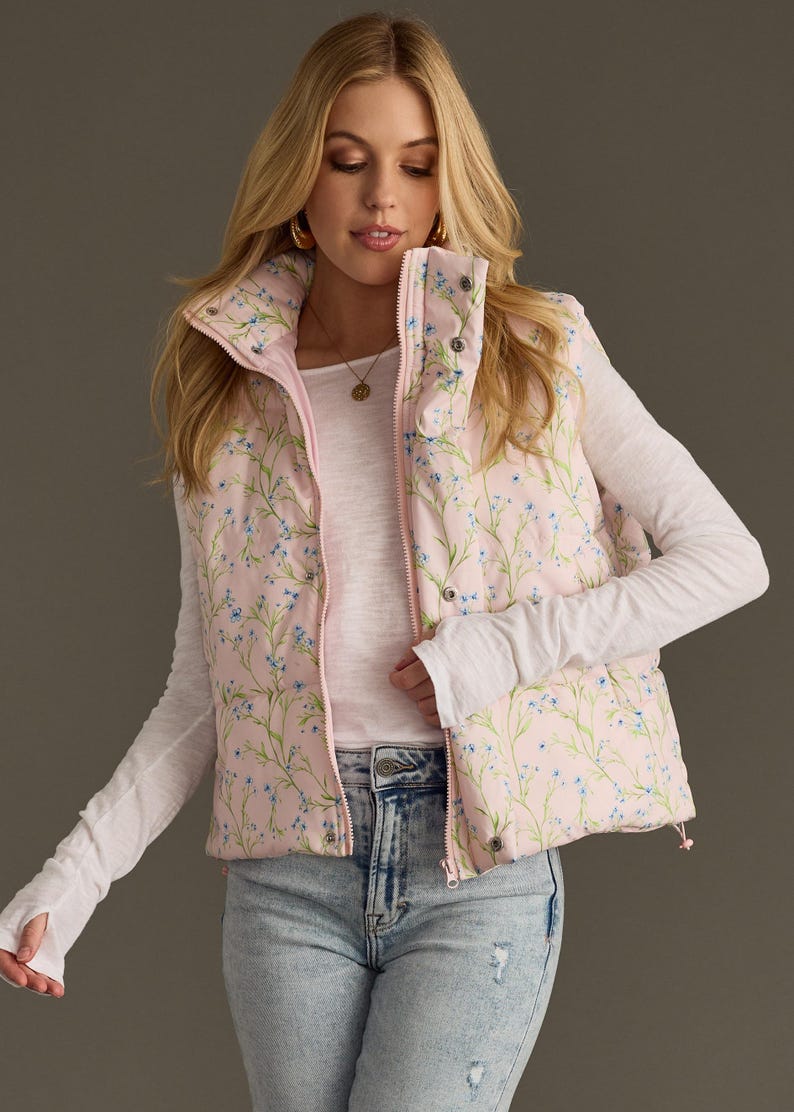 May include: A pink puffer vest with a floral print featuring small blue flowers and green stems. The vest has a zip closure and a stand-up collar.