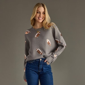 May include: A gray sweater with a pattern of brown and white footballs. The sweater has long sleeves and a ribbed hem and cuffs. The person is wearing blue jeans.