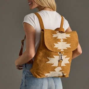 May include: Mustard yellow backpack with a white geometric pattern on the flap. The backpack has adjustable straps and a metal clasp closure. The bag is made of a woven fabric and is worn by a person wearing a white t-shirt and blue jeans.