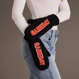 May include: A pair of black knit mittens featuring the word GAMEDAY embroidered in red block letters on the back of each hand. The mittens have a ribbed cuff design and are constructed from a thick, textured knit fabric for warmth and coverage.
