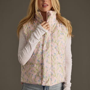 May include: A pink puffer vest with a floral print featuring small blue flowers and green stems. The vest has a zip closure and a stand-up collar.