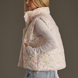 May include: A pink puffer vest with a floral print featuring small blue flowers and green stems. The vest has a zip closure and a stand-up collar.