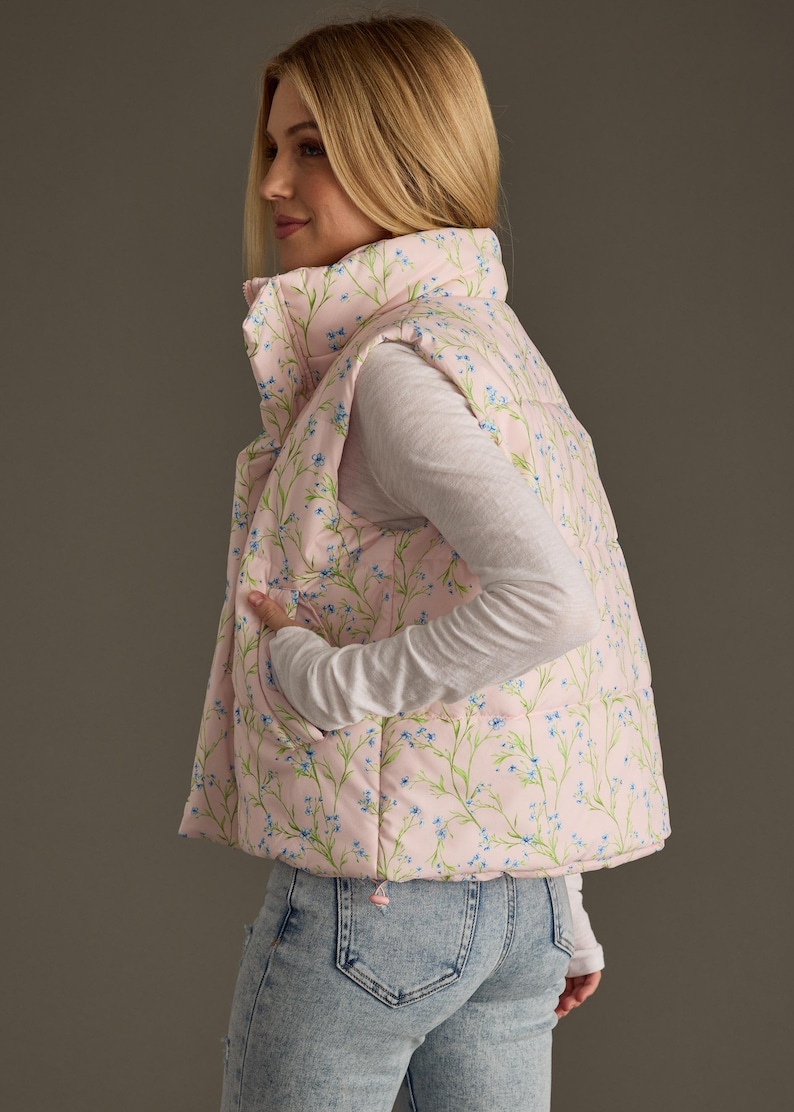 May include: A pink puffer vest with a floral print featuring small blue flowers and green stems. The vest has a zip closure and a stand-up collar.