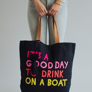 May include: Black tote bag with the embroidered phrase "IT'S A GOOD DAY TO DRINK ON A BOAT" in pink, orange, and yellow. The bag has brown leather handles and a navy tassel. A fun accessory for a day out.