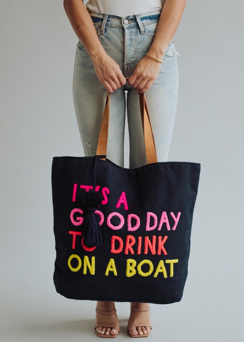 May include: Black tote bag with the embroidered phrase "IT'S A GOOD DAY TO DRINK ON A BOAT" in pink, orange, and yellow. The bag has brown leather handles and a navy tassel. A fun accessory for a day out.