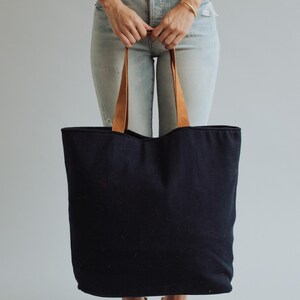 May include: A large black tote bag with brown leather handles. The bag is held by a person wearing light blue jeans and a white top. The tote bag is a simple, classic design.