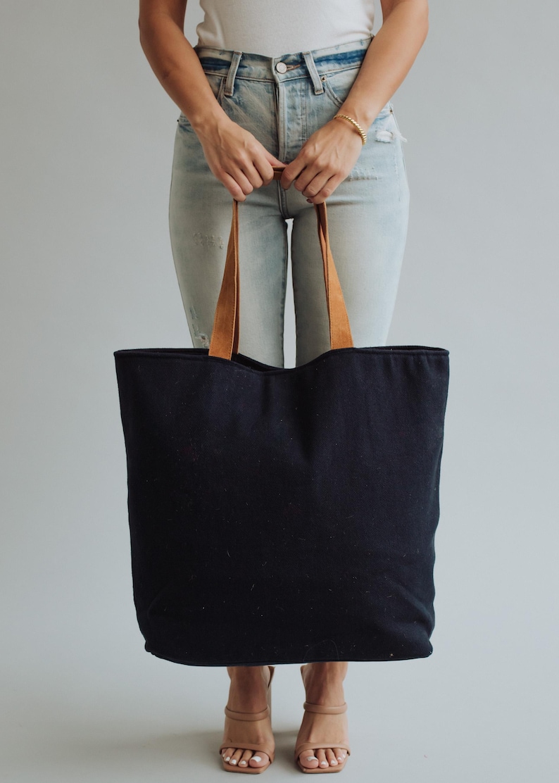 May include: A large black tote bag with brown leather handles. The bag is held by a person wearing light blue jeans and a white top. The tote bag is a simple, classic design.
