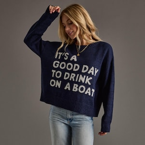 May include: A navy blue long-sleeve knit sweater featuring the white text IT'S A GOOD DAY TO DRINK ON A BOAT across the chest. The sweater has a crew neckline, ribbed cuffs, and a ribbed hem, paired with light blue denim jeans.