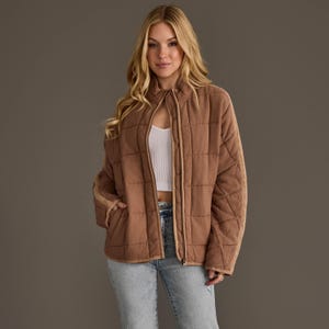 May include: A brown quilted jacket featuring a full-length front zipper, a high collar, and long sleeves. The jacket has a grid-like stitched pattern across the fabric, side pockets, and light-colored trim along the zipper and sleeve edges.
