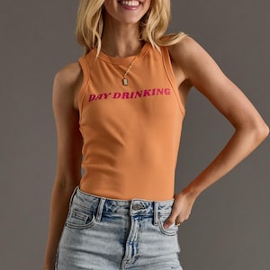 May include: Orange ribbed tank top with the text "DAY DRINKING" in pink. The top is paired with light wash denim jeans. A gold necklace is visible.