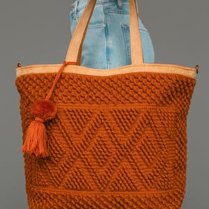 May include: A large burnt orange tote bag featuring a textured bobble knit pattern with a geometric diamond design. The bag includes two light tan shoulder straps and a matching orange yarn tassel attached to the side with a thin cord.