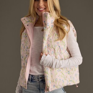 May include: A pink puffer vest with a floral print featuring small blue flowers and green stems. The vest has a zip closure and a stand-up collar.