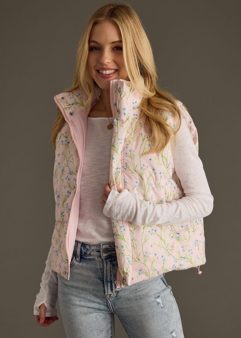 May include: A pink puffer vest with a floral print featuring small blue flowers and green stems. The vest has a zip closure and a stand-up collar.