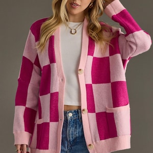 May include: A pink and fuchsia checkerboard pattern cardigan with a light pink trim, buttons, and pockets. The long-sleeved sweater is worn over a white top and blue jeans. The model is wearing a gold necklace and earrings.