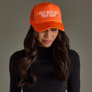 May include: Bright orange trucker hat with white text that reads "I ONLY DATE GUYS THAT HUNT". The hat has a mesh back and a curved brim. The hat is worn by a person with long, dark hair.