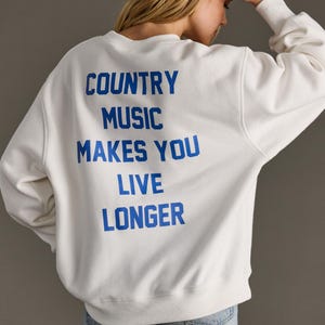 May include: White sweatshirt with the text "COUNTRY MUSIC MAKES YOU LIVE LONGER" in blue lettering. The sweatshirt is a crew neck style with long sleeves. The person in the image is wearing blue jeans.