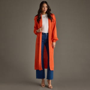 May include: A long, open-front orange knit cardigan with long sleeves and two front pockets, worn over a plain white crew-neck top and dark blue denim flare jeans. The cardigan features a ribbed hem and cuffs, providing a layered look.