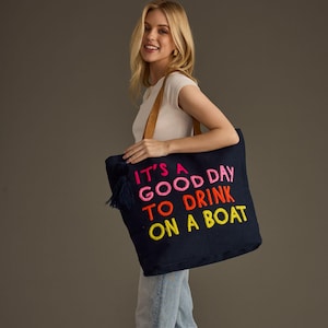 May include: Navy blue tote bag with brown leather straps and a navy tassel. The bag features the embroidered text "IT'S A GOOD DAY TO DRINK ON A BOAT" in pink, orange, and yellow.