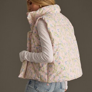 May include: A pink puffer vest with a floral print featuring small blue flowers and green stems. The vest has a high collar and a zip closure.