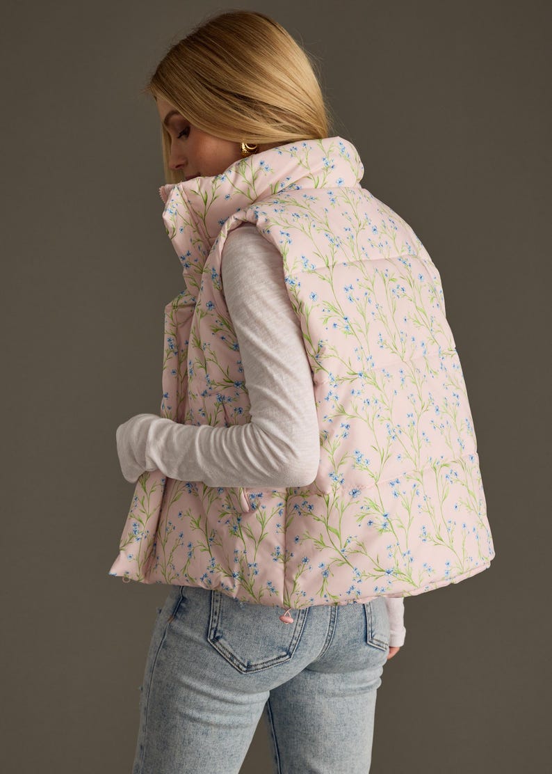 May include: A pink puffer vest with a floral print featuring small blue flowers and green stems. The vest has a high collar and a zip closure.
