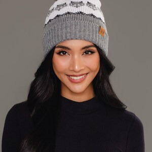 May include: A knit beanie featuring a grey ribbed cuff, a white mid-section with a repeating grey fox pattern, and a brown faux fur pom-pom on top. A small tan rectangular tag is attached to the side of the grey ribbed brim of the winter hat.