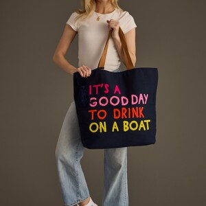 May include: Navy blue tote bag with brown leather handles and the embroidered text "IT'S A GOOD DAY TO DRINK ON A BOAT" in bright pink, orange, and yellow. The bag has a navy tassel and is held by a person.