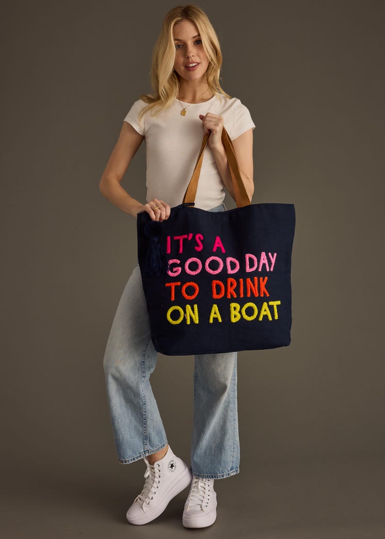 May include: Navy blue tote bag with brown leather handles and the embroidered text "IT'S A GOOD DAY TO DRINK ON A BOAT" in bright pink, orange, and yellow. The bag has a navy tassel and is held by a person.