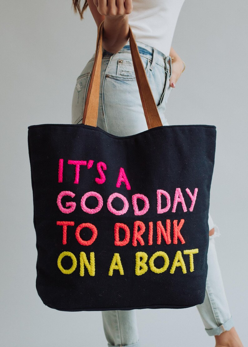 May include: Black tote bag with brown leather handles. The bag features embroidered text in pink, orange, and yellow that reads "IT'S A GOOD DAY TO DRINK ON A BOAT". The bag is held by a person wearing light blue jeans.