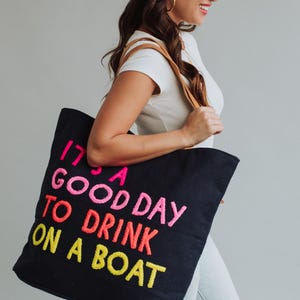 May include: Black tote bag with the embroidered phrase "IT'S A GOOD DAY TO DRINK ON A BOAT" in pink, orange, and yellow. The bag has brown leather straps and is carried by a person wearing a white shirt and jeans.