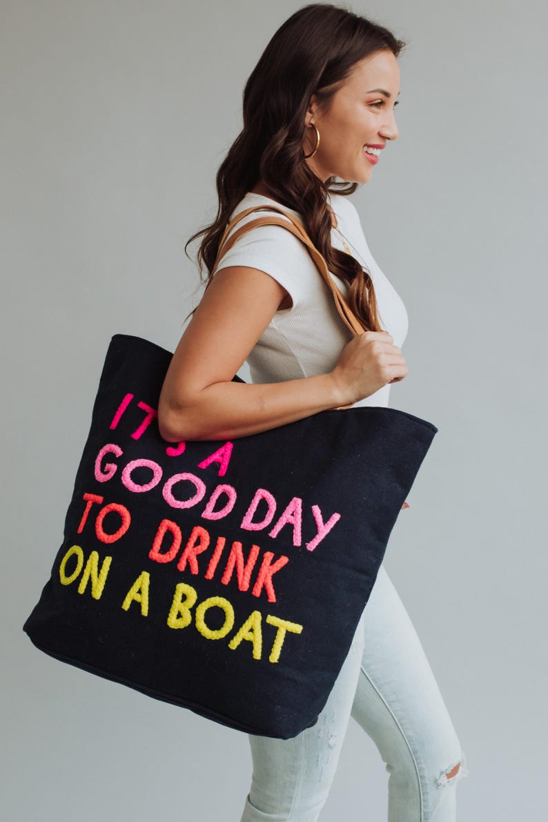 May include: Black tote bag with the embroidered phrase "IT'S A GOOD DAY TO DRINK ON A BOAT" in pink, orange, and yellow. The bag has brown leather straps and is carried by a person wearing a white shirt and jeans.