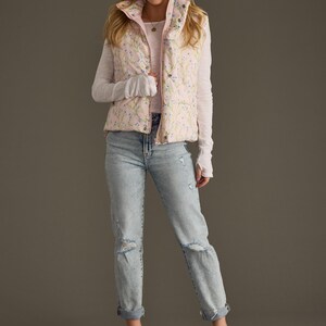 May include: A pink floral print puffer vest worn over a white long-sleeved shirt and light blue jeans. The person is wearing light blue sandals.